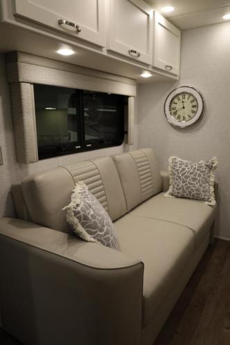  Inside the 2025 Newmar Bay Star Sport, the living area is a testament to modern comfort and style, with a beige sofa, decorative cushions, and thoughtful touches like a chic clock and ample storage, set against the backdrop of an exterior view.